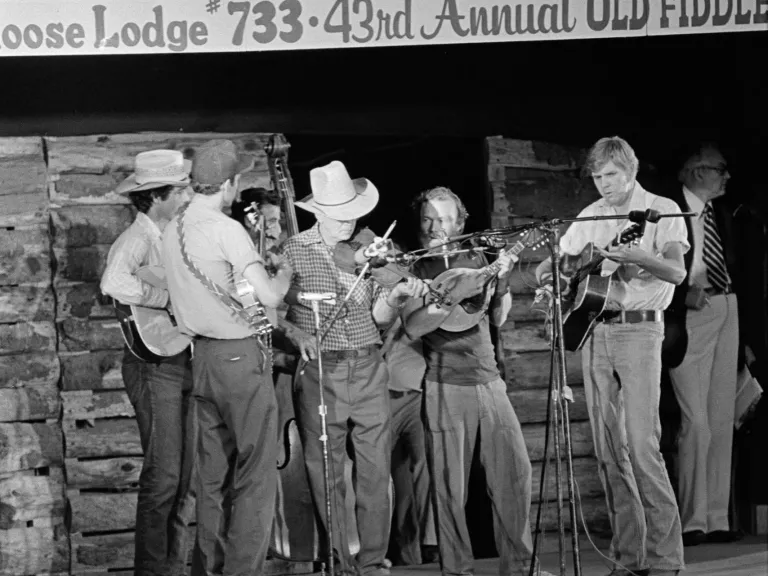 A group of musicians on a stage play fiddles. Some wear boots and cowboy hats.