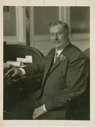 A sepia photograph of James E. Crass in a suit sitting at a roll-top desk