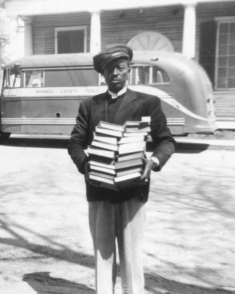 Black and white photograph of an African American man holding two stacks of books in front of the Virginia State Library Bookmobile