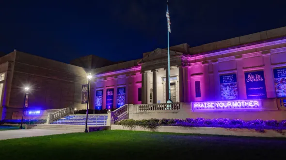 The front of the museum at night illuminated by two neon signs spelling out "Praise Your Mother / Ama A La Mama" in blue and purple.