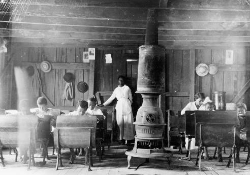 Black and white photograph of the interior of a one room African American schoolhouse, showing a coal powered chimney in the center of the room with students working at desks around it.  