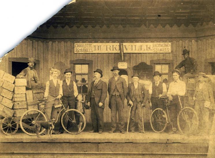 Burkeville Train Station (VHS accession number: 1999.160.969). Black and white albumen silver print shows the Burkeville Depot. A sign hanging on the wall reads: Charlotte 228 6/10 miles, Richmond 53 4/10 miles. On the left, there are wooden crates piled upon a cart. One man sits on that cart. Next to him, a two men and a child stand with a bicycle and a dog. The child is holding the hand of a third man, who is wearing a suit. Two other men wearing suits are standing next to him. The man on the right is elderly and holds two canes. Next to the elderly man, two other men are standing by another bicycle. An African American man is sitting on top of some crates to the right above the man to the furthest right, who is holding a newspaper or something in his hands. A chunk of the photograph has been broken off in the top left corner. Burkeville Train Station
