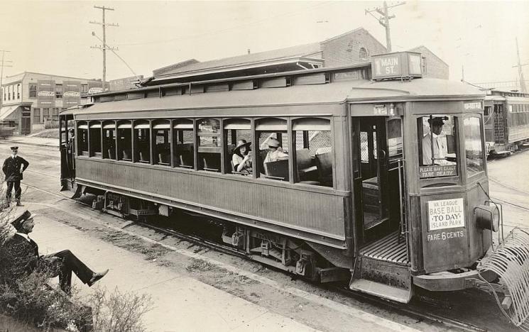 Streetcar, Richmond (VHS accession number: 2000.186.586.b) Photographic print, Streetcar, Richmond, Virginia. Glossy black and white photographic print shows a streetcar parked next to a stop on the Main Street line in Richmond, Virginia. Sign on the front of the streetcar reads "VA. LEAGUE / BASEBALL / TODAY / ISLAND PARK." A sign painted on the streetcar indicates that the fare is six cents. Two women are sitting on the streetcar. A driver and conductor are also on board the streetcar. Two additional conductors, one seated and one standing, are to the left. Brick buildings and another streetcar are in the background. 20.32 cm x 25.40 cm. Streetcar, Richmond