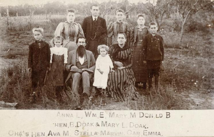 Doak Family (VHS accession number: 2001.345.1). Photograph, Doak Family. Black and white image measuring 4" x 6 1/4" mounted on decorative board. Photograph shows family group consisting of a mother and father, four young children and four adult children. The mother wears a striped dress and the father has a full dark beard. They sit and stand in a formal pose in an austere landscape. There is an inscription identifiying the subjects in the photograph, but it is difficult to read. Names listed are follows: Anna L., Wm. E., Mary M., Donald B., Hen, B. Doak & Mary L. Doak, Cha's Hen, Ava M., Stella Marvin, Car., Emma. Doak Family