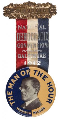 A red, white, and blue ribbon with the words, “NATIONAL DEMOCRATIC CONVENTION / BALTIMORE / 1912”, above a blue and yellow button with a black and white photograph of Woodrow Wilson placed in the middle with the text, “THE MAN OF THE HOUR / WOODROW WILSON.” 