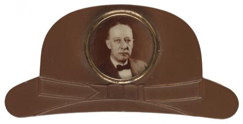A lapel pin in the shape of a brown derby hat with central photo of Al Smith