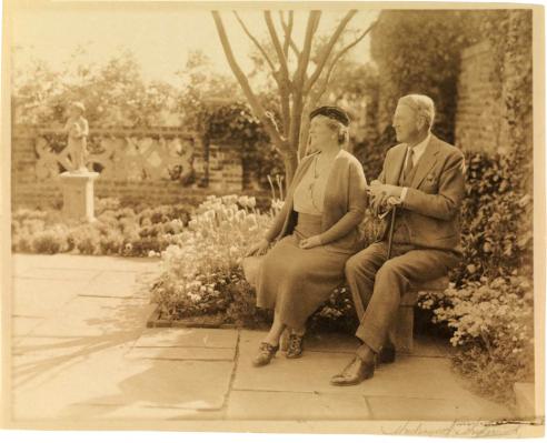A vintage sepia colored photograph of Virginia and Alexander Weddell sitting on a bench in the Four Seasons Garden surrounded by flowers