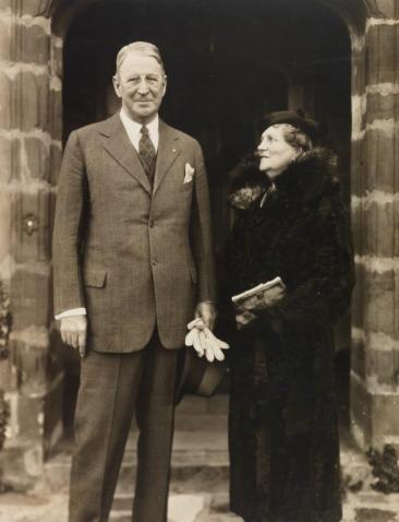 Photograph of the Weddells holding hands standing in an open walkway. Virginia Weddell can be seen facing and smiling at Alexander Weddell as he looks towards the camera. 